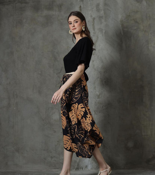 A woman poses against a textured gray background, wearing a black top and flowy summer pants with a V hem, adorned with a bold orange leaf pattern. She accessorizes with large earrings and strappy sandals, looking over her shoulder.