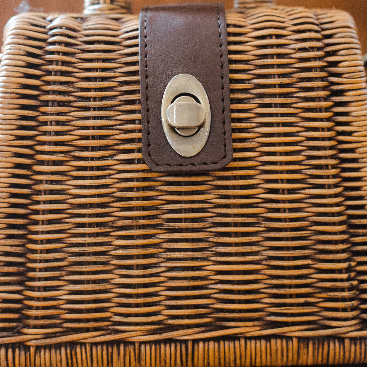 A close-up of a brown woven handbag with a leather strap and a metallic twist lock. The texture of the weave is detailed, showcasing the craftsmanship. The lock is centered on the strap.