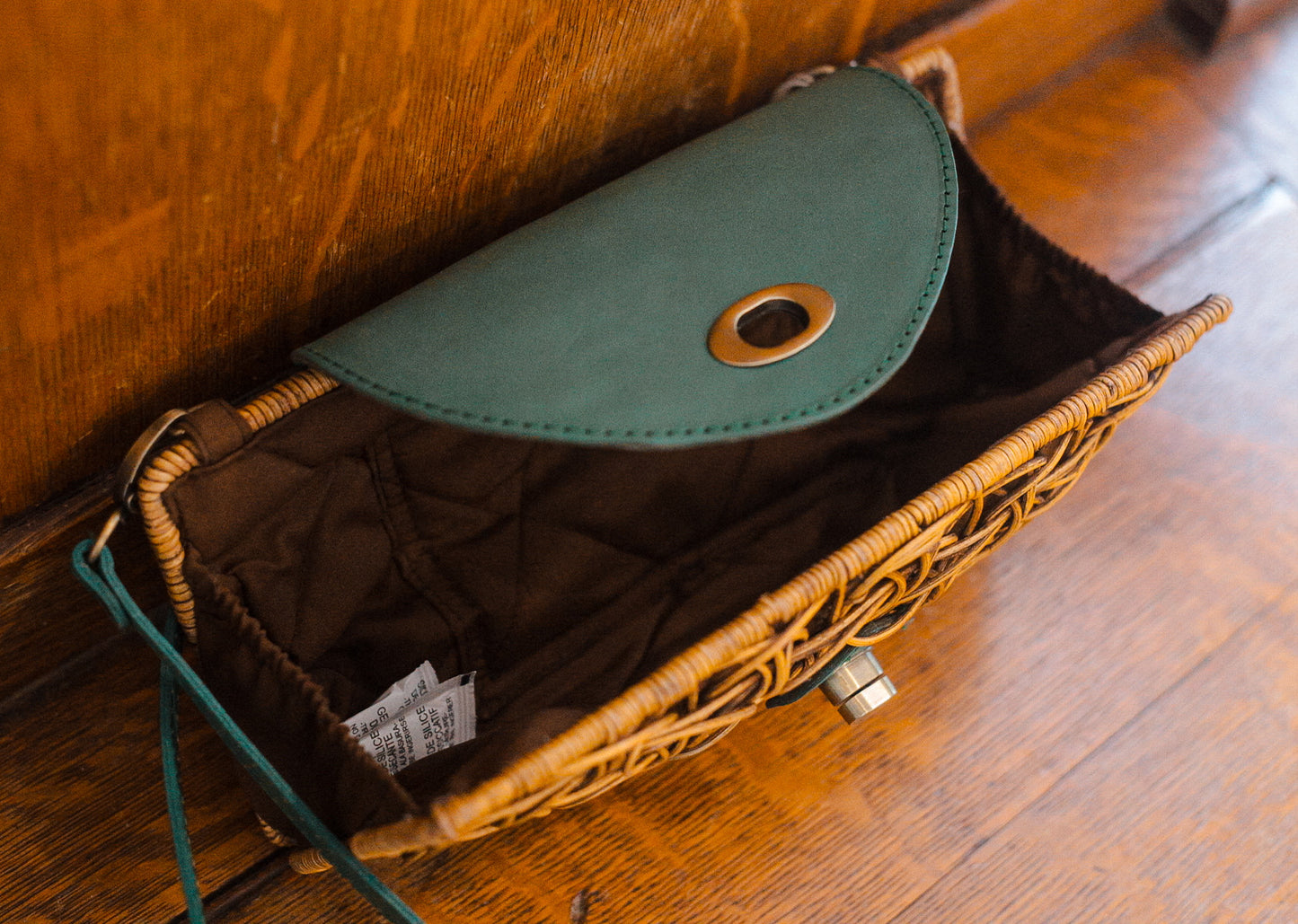 A small woven rattan purse with a green flap is open, revealing a dark brown interior. It rests on a wooden surface, with visible metal fittings and a tag inside.