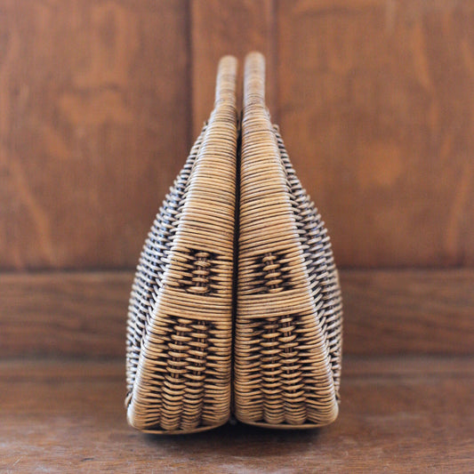 A woven wicker handbag standing upright on a wooden surface against a wooden background. The bag has a symmetrical design with a tightly knit pattern, featuring natural brown tones.