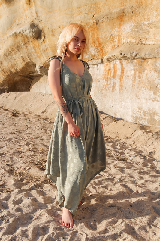 A woman with blonde hair stands barefoot on a sandy beach, wearing a green patterned long jumpsuit. She poses confidently against the backdrop of a sunlit, rocky cliff, creating a natural and serene atmosphere.