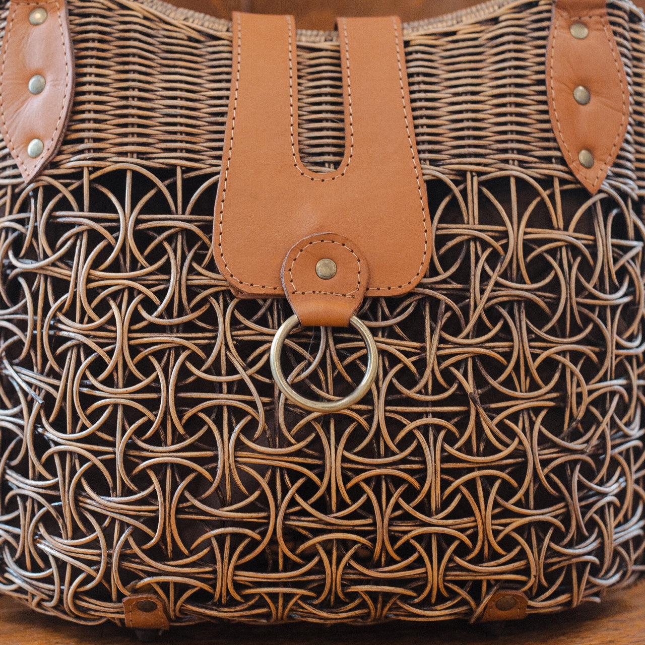 Close-up of a wicker basket with an intricate interlaced pattern. It features tan leather accents and a brass ring at the center. The texture and craftsmanship are prominently displayed, highlighting the baskets detailed design.