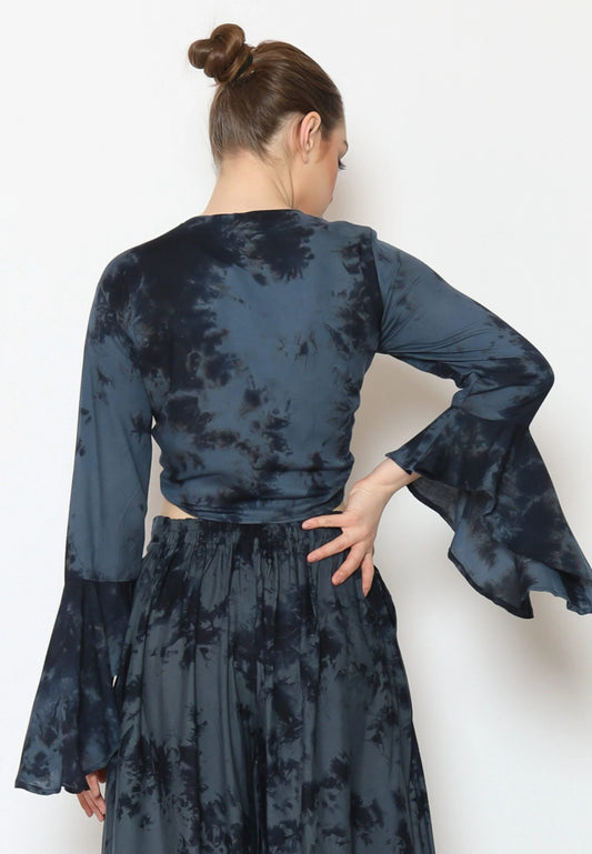 A person with hair in a bun is wearing a dark blue and black boho top featuring flared sleeves and a matching skirt. The person is facing away, standing against a plain white background.