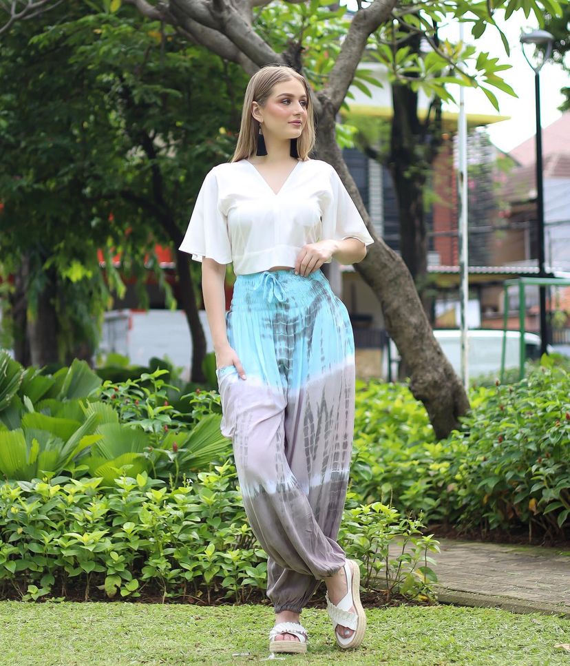 A woman stands in a garden wearing a white top and blue-gray pants for women, with trees and greenery in the background.