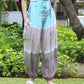 Person wearing blue and gray tie-dye pants and white sandals stands on grass with greenery in the background.