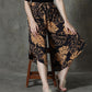 A person sits gracefully on a wooden stool, donning black and brown leaf-patterned summer pants with a stylish V hem. Their beige sandals rest comfortably on the floor, set against a textured gray wall.