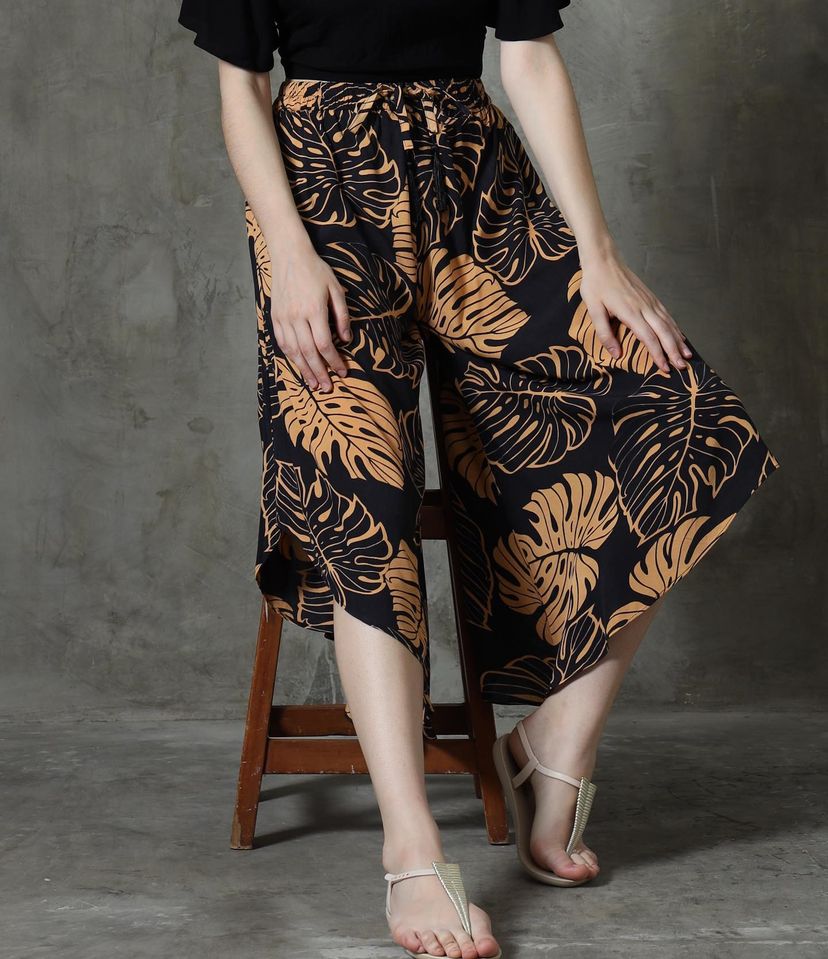 A person sits gracefully on a wooden stool, donning black and brown leaf-patterned summer pants with a stylish V hem. Their beige sandals rest comfortably on the floor, set against a textured gray wall.