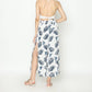A woman stands with her back to the camera, wearing a halter-neck dress adorned with a tropical palm print. The sexy white fabric, decorated with blue leaves, features a side slit. Her shoulder-length blonde hair flows freely as she pairs the look effortlessly with sandals.