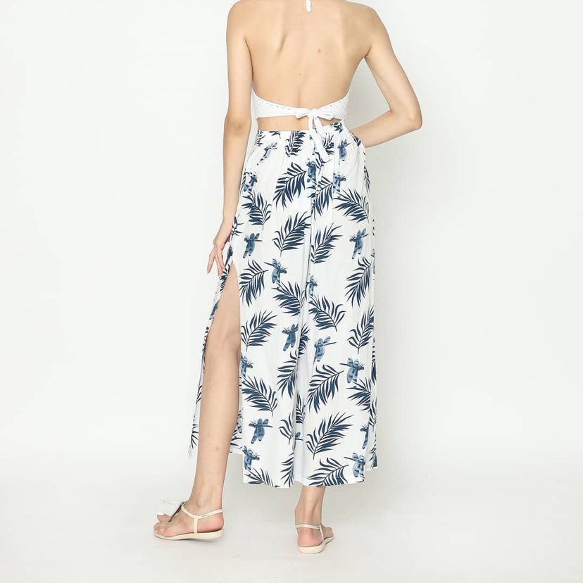 A woman stands with her back to the camera, wearing a halter-neck dress adorned with a tropical palm print. The sexy white fabric, decorated with blue leaves, features a side slit. Her shoulder-length blonde hair flows freely as she pairs the look effortlessly with sandals.