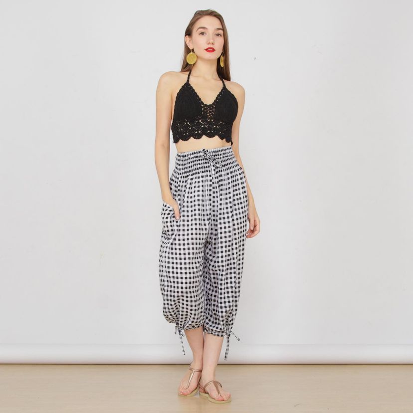 A woman stands against a white background, wearing a black lace halter top and chic high-waisted, 3/4 length checkered pants. She has long hair, bold red lipstick, gold hoop earrings, and is barefoot.