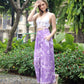 A woman stands outdoors, wearing a white crochet crop top and flowing purple tie-dye pants. Shes holding her hair and smiling slightly. The background features greenery and buildings.