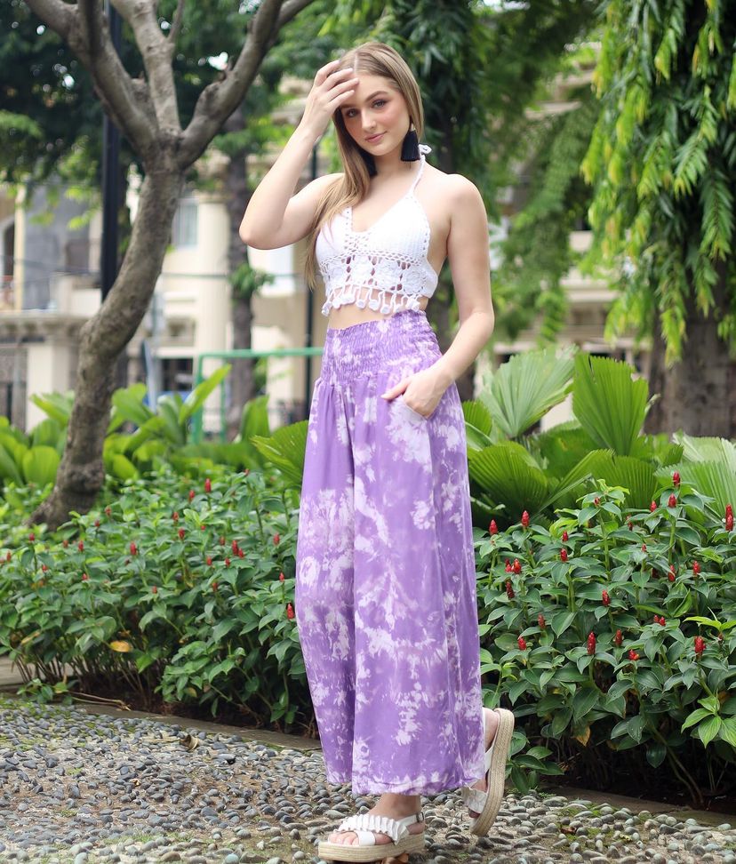 A woman stands outdoors, wearing a white crochet crop top and flowing purple tie-dye pants. Shes holding her hair and smiling slightly. The background features greenery and buildings.