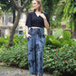 A woman in a black halter top and blue boho flare pants poses outdoors on a pebble path. She is wearing white sandals, surrounded by lush green plants and trees.