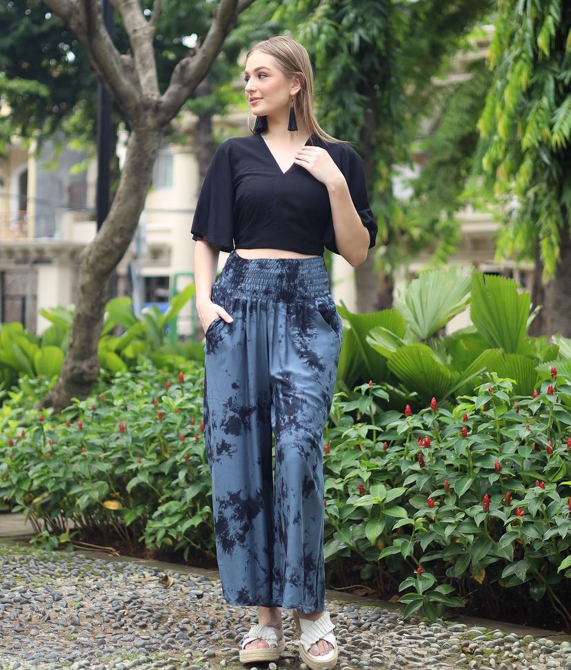 A woman in a black halter top and blue boho flare pants poses outdoors on a pebble path. She is wearing white sandals, surrounded by lush green plants and trees.