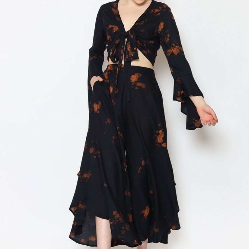 A person in a black tie-front top with long flared sleeves and matching wide-leg pants featuring vibrant orange tie-dye patterns stands gracefully against a plain white background, evoking the charm of a ruffle skirt.