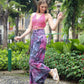 A woman in a pink halter top and boho flare pants poses joyfully on a stone path in a leafy park. She wears platform sandals and is surrounded by greenery and flowering plants.
