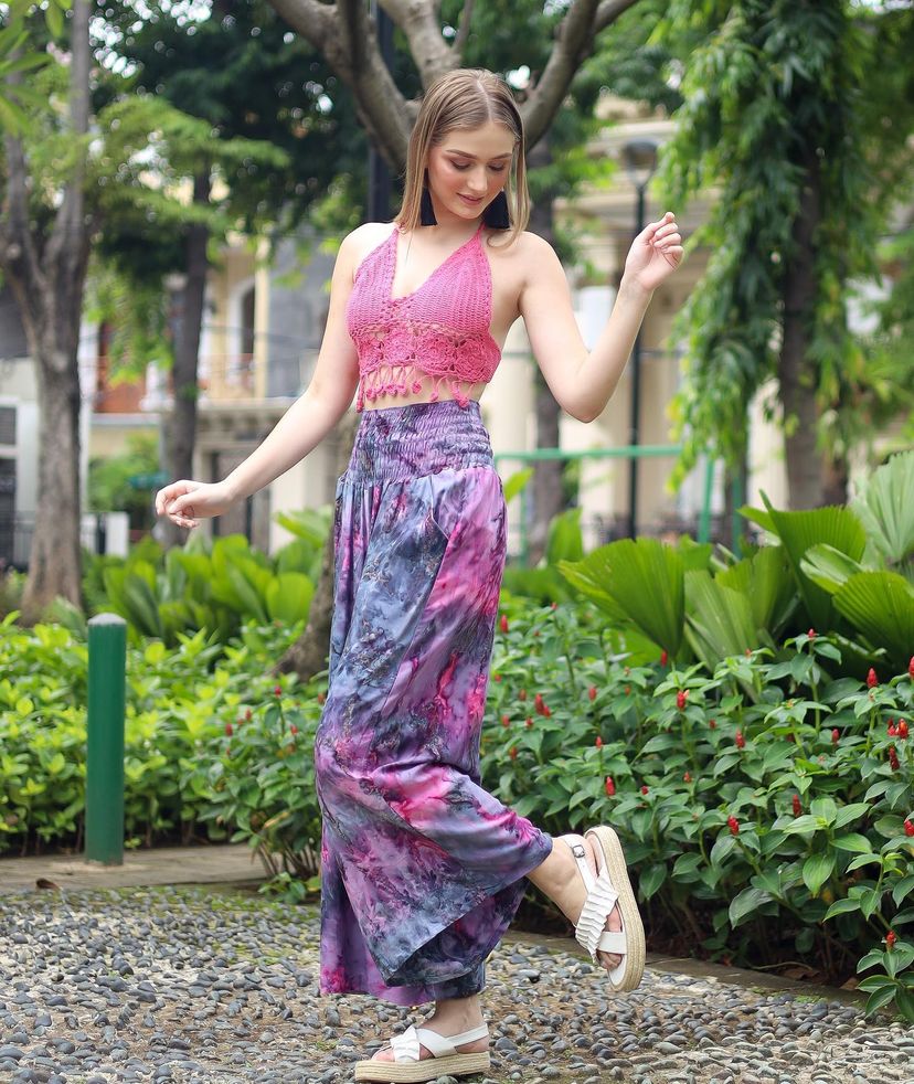A woman in a pink halter top and boho flare pants poses joyfully on a stone path in a leafy park. She wears platform sandals and is surrounded by greenery and flowering plants.