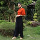 A person in an orange top and black yoga pants stands in a garden with lush greenery, smiling and looking down. They are wearing sandals, and a gazebo is visible in the background.