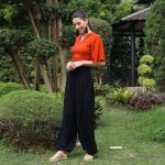 A person in an orange top and black yoga pants stands in a garden with lush greenery, smiling and looking down. They are wearing sandals, and a gazebo is visible in the background.
