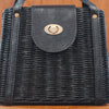 A close-up of a black woven handbag with a leather strap and a metallic twist lock. The texture of the weave is detailed, showcasing the craftsmanship. The lock is centered on the strap.