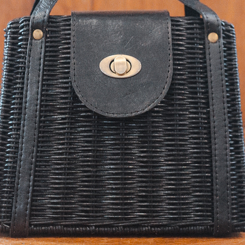 A close-up of a black woven handbag with a leather strap and a metallic twist lock. The texture of the weave is detailed, showcasing the craftsmanship. The lock is centered on the strap.