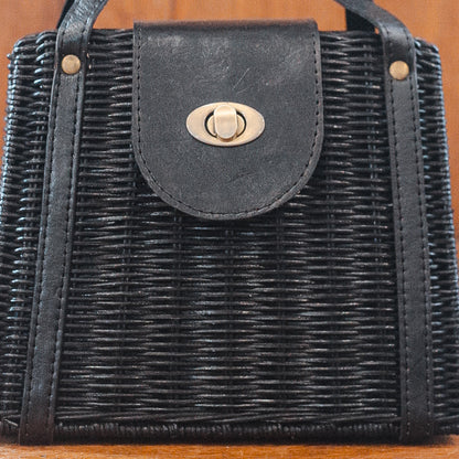A close-up of a black woven handbag with a leather strap and a metallic twist lock. The texture of the weave is detailed, showcasing the craftsmanship. The lock is centered on the strap.