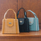 A trio of small woven handbags with leather accents, including charming small rattan purses, are elegantly displayed on a wooden surface. The collection boasts three colors: beige with a tan flap, black with matching details, and light blue. Each features a circular clasp and handle for style and function.