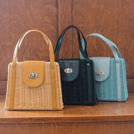 A trio of small woven handbags with leather accents, including charming small rattan purses, are elegantly displayed on a wooden surface. The collection boasts three colors: beige with a tan flap, black with matching details, and light blue. Each features a circular clasp and handle for style and function.