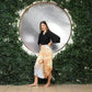 A woman stands confidently in front of a circular mirror framed by lush green leaves. She wears a black blouse with long sleeves and short pants with a v hem, paired with sandals. Her hair is shoulder-length, and she smiles slightly.