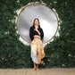 A woman stands confidently against a circular mirror backdrop framed by lush green foliage. She wears a black top and an asymmetrical, multicolored skirt over short pants with a v hem, her hands held gracefully in front.