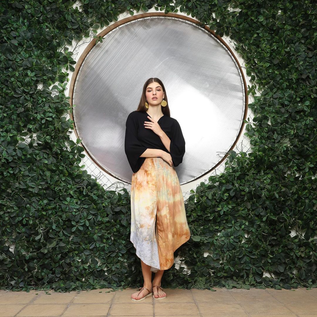 A woman stands confidently against a circular mirror backdrop framed by lush green foliage. She wears a black top and an asymmetrical, multicolored skirt over short pants with a v hem, her hands held gracefully in front.
