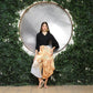 A woman stands confidently in front of a circular mirror surrounded by greenery. Sporting a black top and vibrant, wide-legged pants with a unique V hem, she completes her look with sandals. Her hand rests on her hip, accentuating her long, straight hair in this serene outdoor setting.