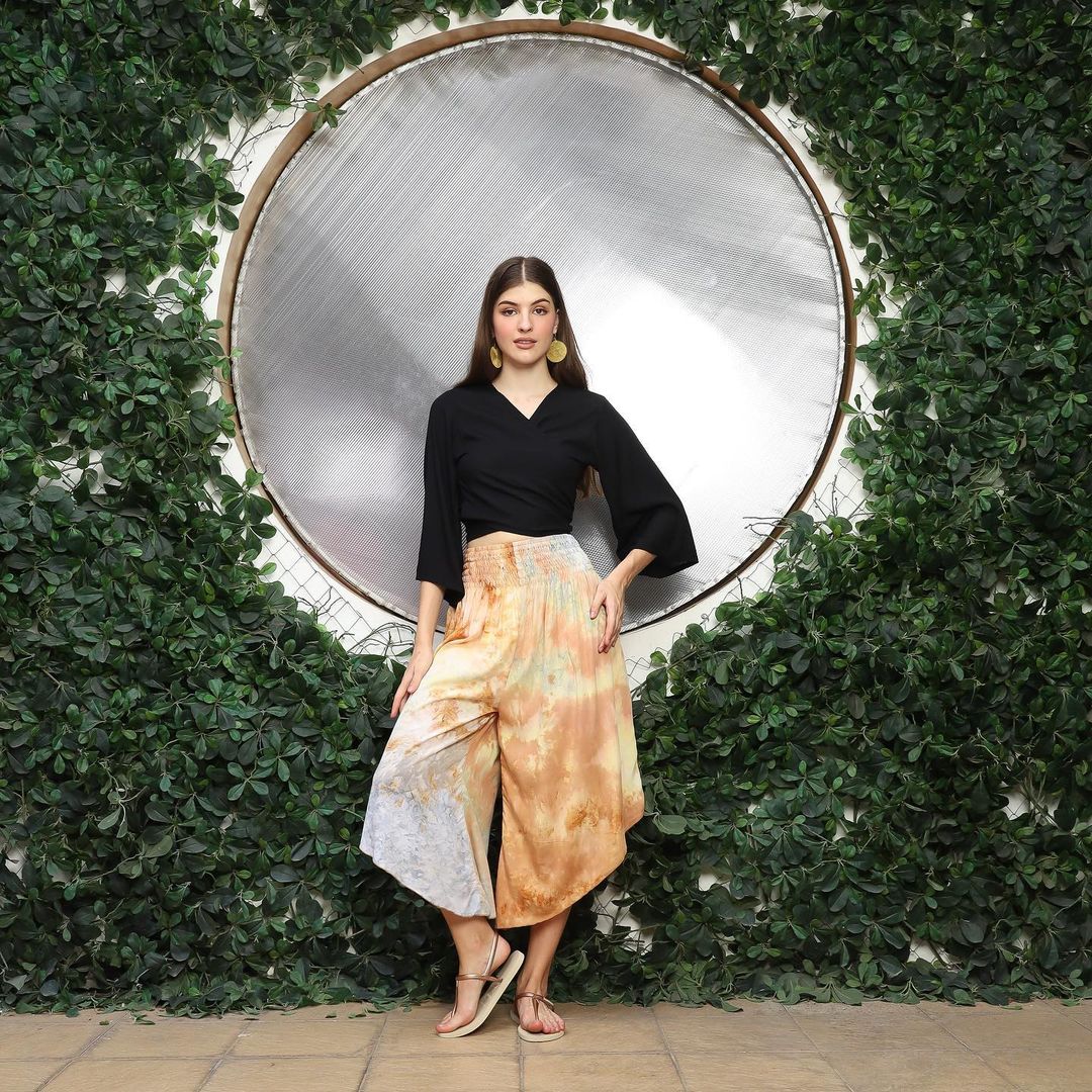 A woman stands confidently in front of a circular mirror surrounded by greenery. Sporting a black top and vibrant, wide-legged pants with a unique V hem, she completes her look with sandals. Her hand rests on her hip, accentuating her long, straight hair in this serene outdoor setting.