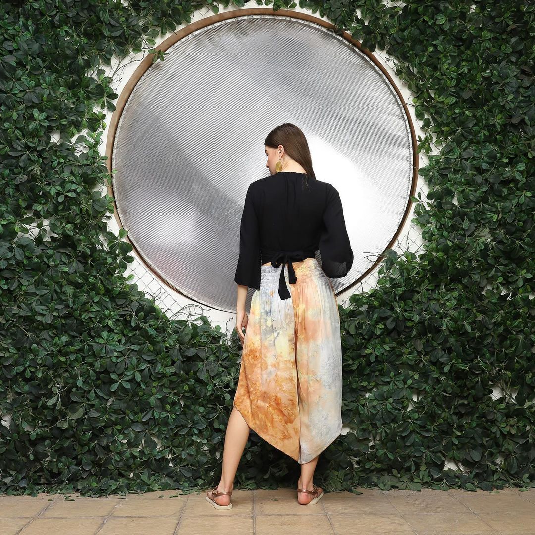 A woman with long hair stands in front of a large circular mirror. She is wearing a black top and a flowy skirt with orange and blue patterns, while in the background, lush green foliage frames the scene beautifully.