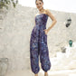 A woman stands outdoors against a light stone wall, wearing a purple and blue patterned strapless romper. Her hair is pulled back as she smiles, one hand resting on her hip. Decorative vases are artfully placed on the steps nearby.