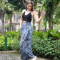 A woman poses outdoors on a pebble path, wearing a black halter top and blue tie-dye boho flare pants, paired with white sandals. She stands among lush greenery and trees, with residential buildings in the background.