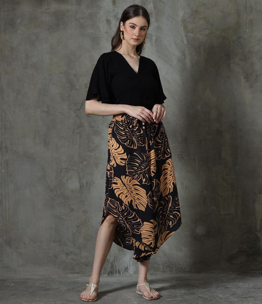 A woman stands against a textured gray background, wearing a black top and skirt adorned with large tan leaf patterns. Her white sandals echo the summer vibe while hoop earrings add elegance, as her hands rest near her skirt pockets, reminiscent of laid-back days in summer pants with V hem.