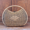 A woven rattan handbag with a rounded shape, featuring a short curved handle. This stylish purse has a crisscross pattern in the center and a wooden background, exuding natural elegance.