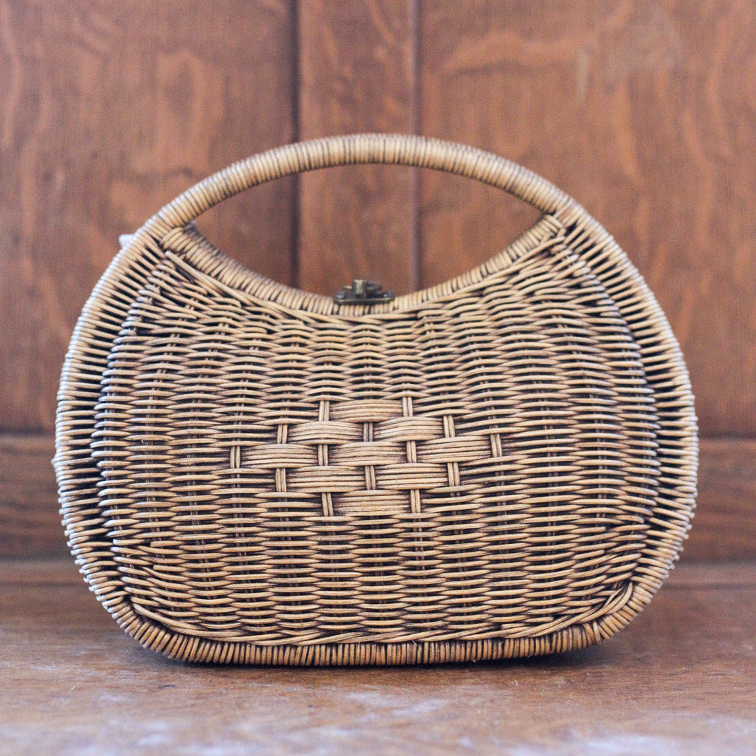 A woven rattan handbag with a rounded shape, featuring a short curved handle. This stylish purse has a crisscross pattern in the center and a wooden background, exuding natural elegance.
