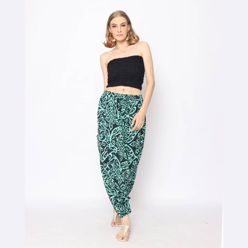 A woman stands against a white background wearing a black strapless top and high-waisted teal pants with a black ornate pattern. She is in sandals and has mid-length light hair. Her pose is relaxed with one hand on her hip.