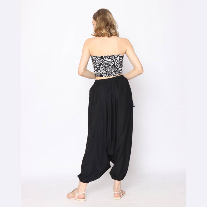 A person with light brown hair stands facing away from the camera, wearing a black and white patterned tube top and loose black harem pants. They are barefoot on a plain white background.