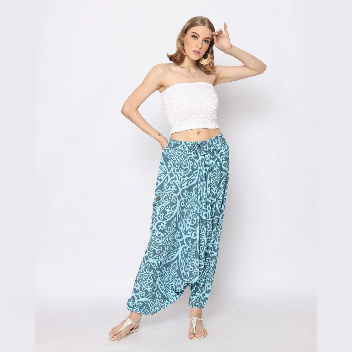 Woman posing against a white background, wearing a strapless white top and loose, patterned blue harem pants for women. She has shoulder-length hair and stands with one hand on her hip and the other raised near her head.