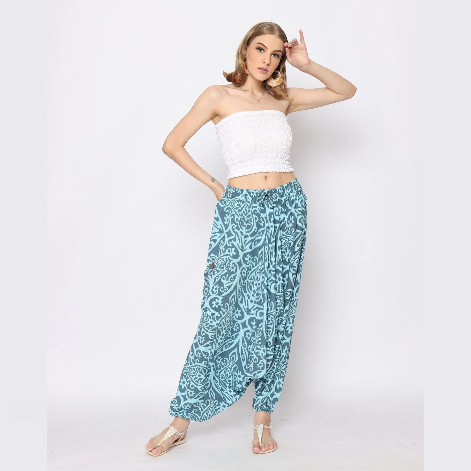 Woman posing against a white background, wearing a strapless white top and loose, patterned blue harem pants for women. She has shoulder-length hair and stands with one hand on her hip and the other raised near her head.
