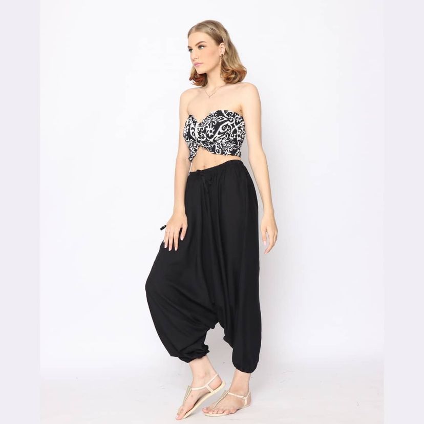 A woman stands against a plain white background wearing a black and white patterned crop top and loose black pants. She has light brown hair and is wearing beige sandals, looking to the side with a relaxed posture.