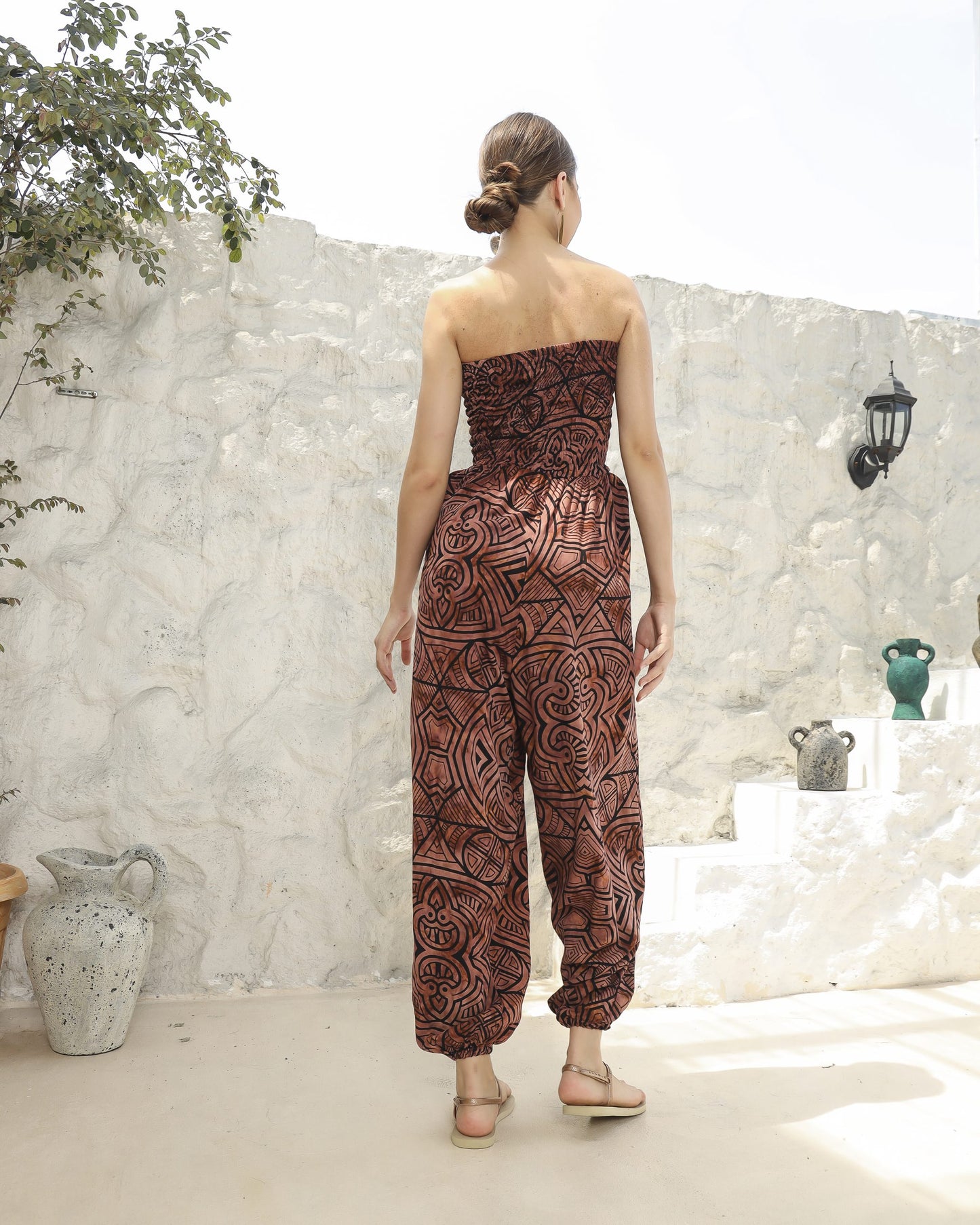 A woman with her back turned stands outdoors in a strapless romper with a bold pattern. She is on a light-colored patio with white stone walls and potted plants. The weather appears sunny.