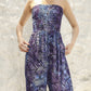 A person wearing a strapless, patterned jumpsuit with geometric designs stands against a textured, light-colored wall. The outfit features shades of blue, purple, and black with intricate swirling and angular patterns.