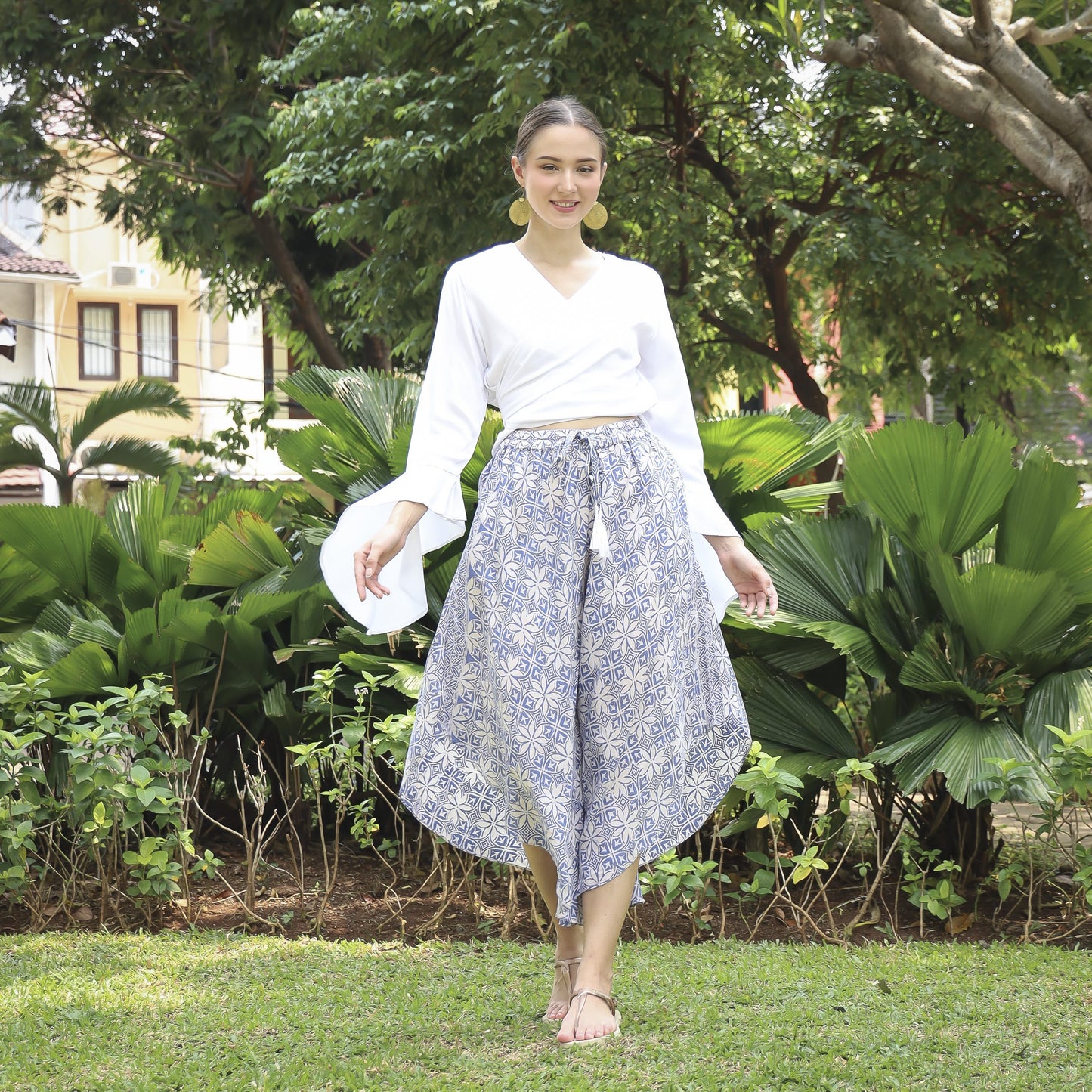 A woman in a white blouse and hand stamped batik pants walks on grass surrounded by lush greenery. She carries a white hat and smiles, wearing sandals, with trees and a building in the background.