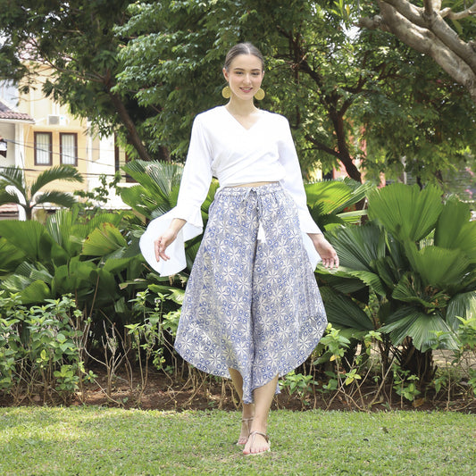 A woman in a white blouse and hand stamped batik pants walks on grass surrounded by lush greenery. She carries a white hat and smiles, wearing sandals, with trees and a building in the background.