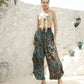 A woman stands outdoors on a sunny day, leaning against a white stone wall. She is wearing a beige crop top and flowing, boho harem pants in shades of teal and brown, with sandals. Potted plants are in the background.