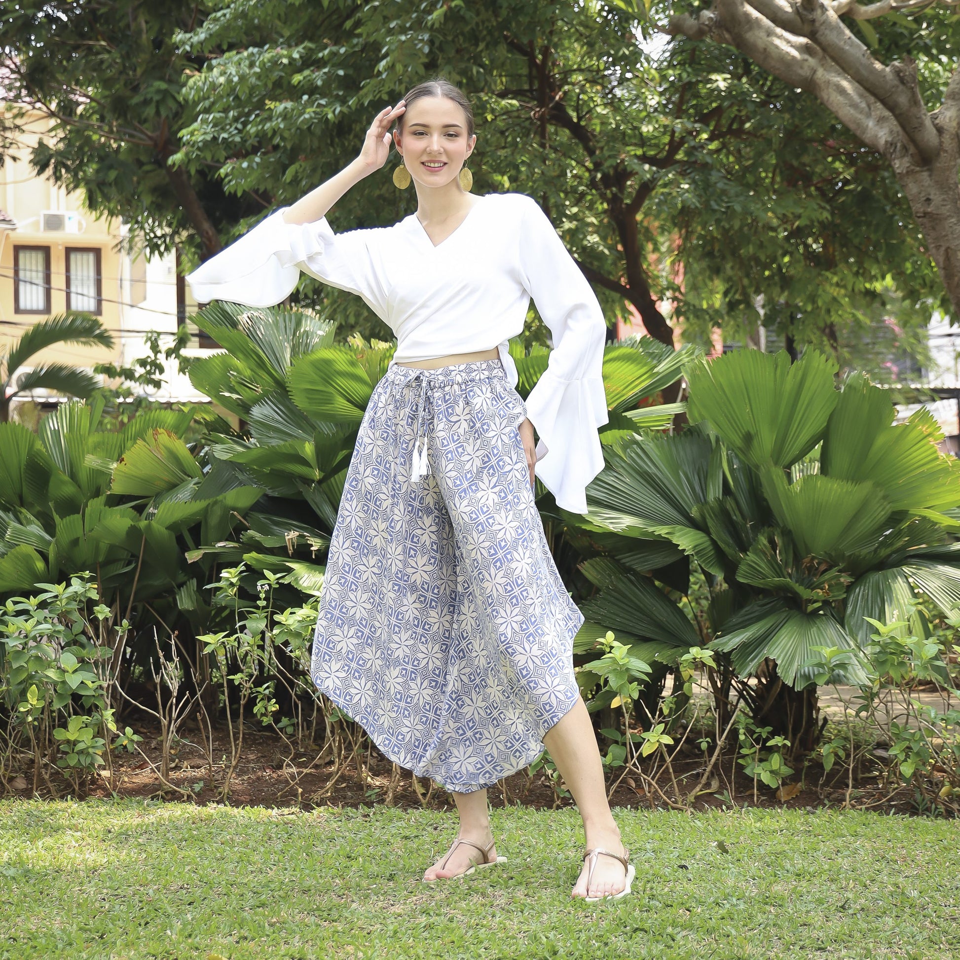 A person stands outdoors on grass, posing with one hand on their head. They are wearing a white blouse with flared sleeves and wide-legged, hand-stamped batik pants in blue and white. Lush green foliage and trees provide a backdrop.
